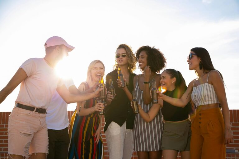 Group of joyful multiracial friends in summer outfits clinking beer bottles and laughing while gathering together on sunny rooftop
